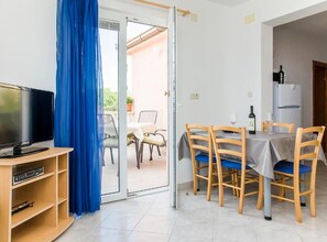 Ein Zimmer mit Fernseher, Esstisch mit Stühlen, Kühlschrank und einem blauen Vorhang.