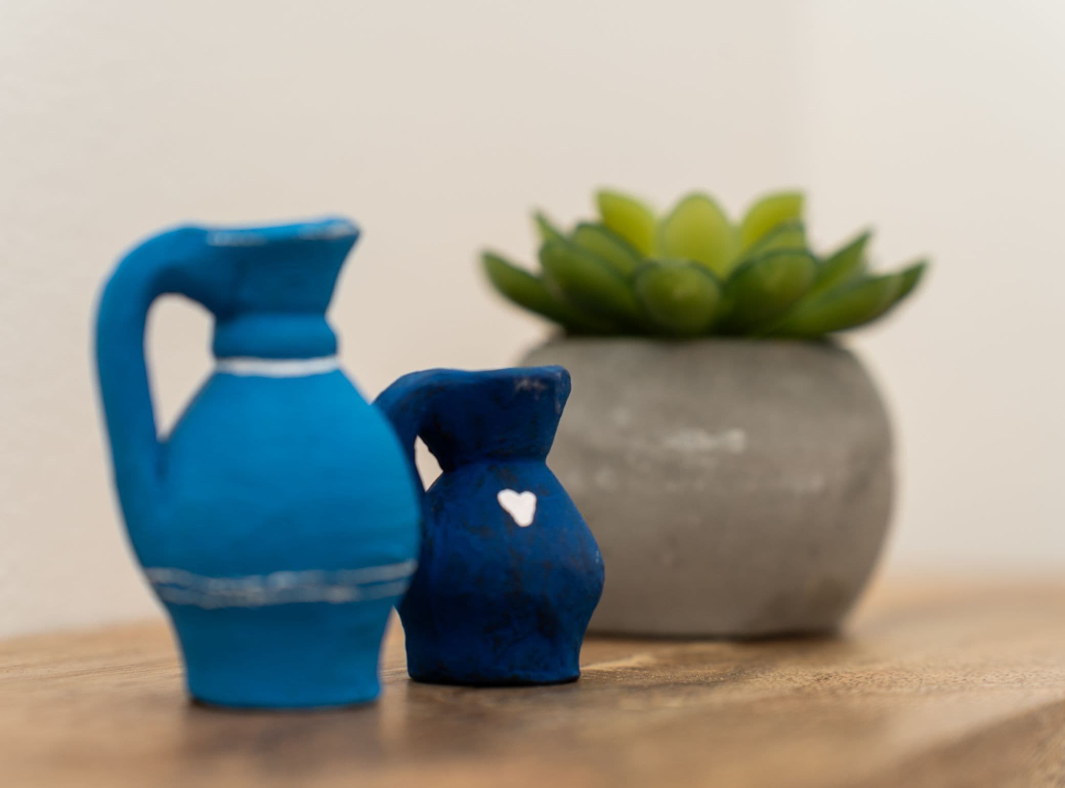 A small blue pitcher and a smaller blue container with a heart design, placed on a wooden surface with a green plant in a grey pot in the background.