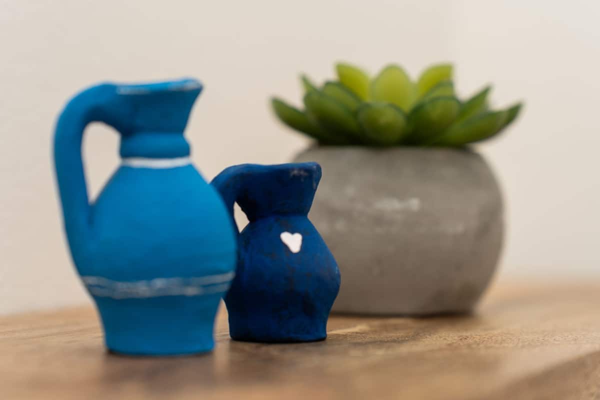 A small blue pitcher and a smaller blue container with a heart design, placed on a wooden surface with a green plant in a grey pot in the background.