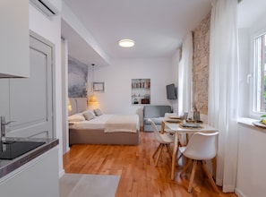 A modern room with a bed, a dining table with chairs, a kitchen area, and a stone wall. - Apartments Nonna (Komiza)