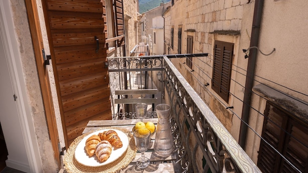 A narrow balcony with a table set for breakfast, including croissants and a glass of water, overlooking a historic street with stone buildings and a mountain in the background. - Apartments Nonna (Komiza)