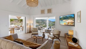 Uma sala de estar ampla com sofá, poltronas, mesa de centro e televisão. Janelas grandes oferecem vista para o exterior.