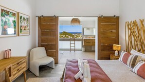 Quarto com cama, cadeira, escrivaninha e vista para a praia através de uma porta aberta.
