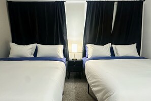 Ein Hotelzimmer mit zwei Betten, jeweils mit weißer Bettwäsche und blauen Akzenten, einem Nachttisch mit Lampe und schwarzen Vorhängen.