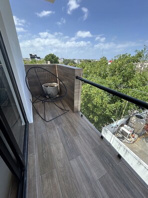 Uma varanda com cadeira, corrimão e vista para árvores e edifícios.