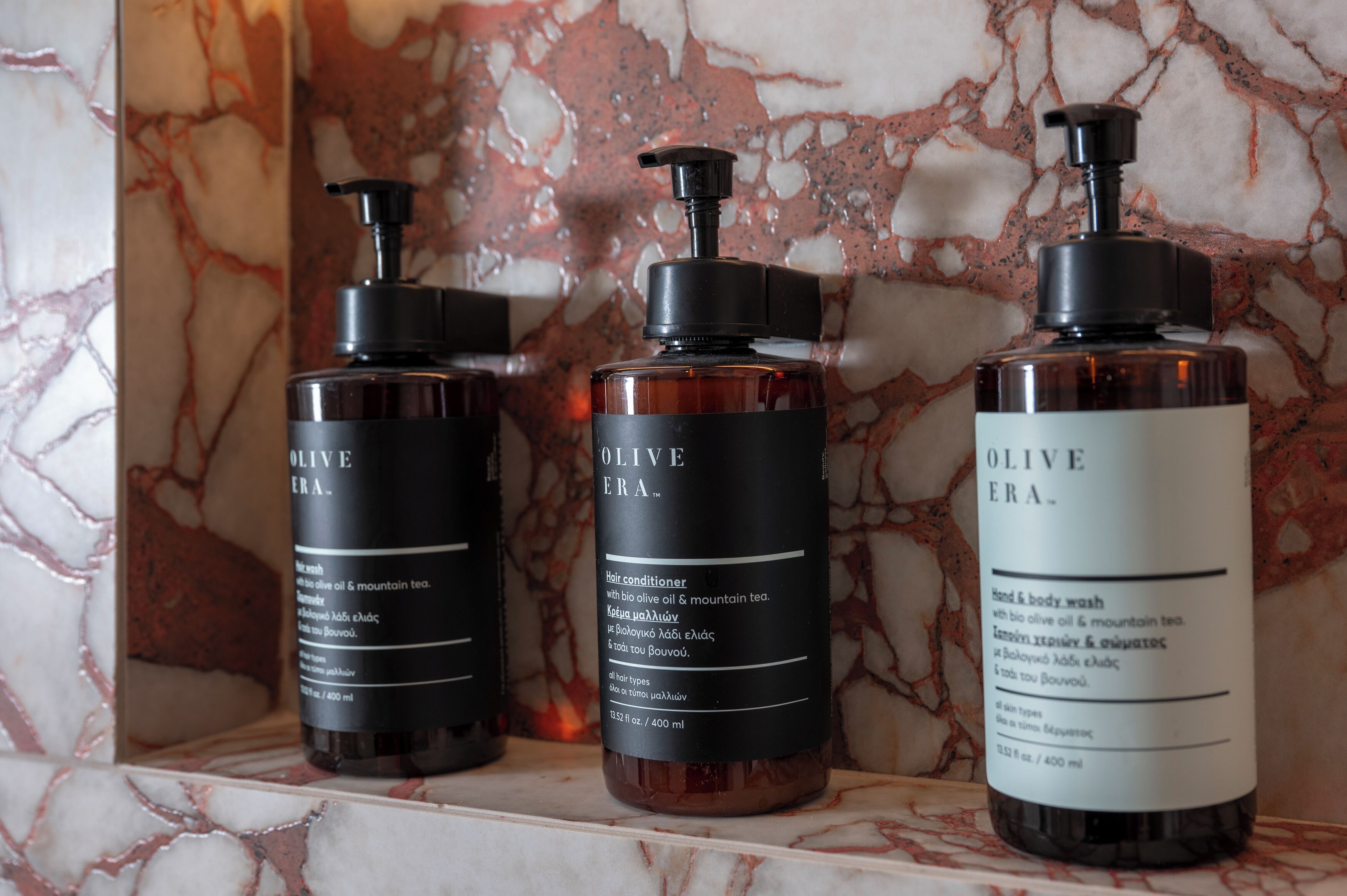 Tres botellas de acondicionador para cabello Olive Era sobre un estante de mármol.