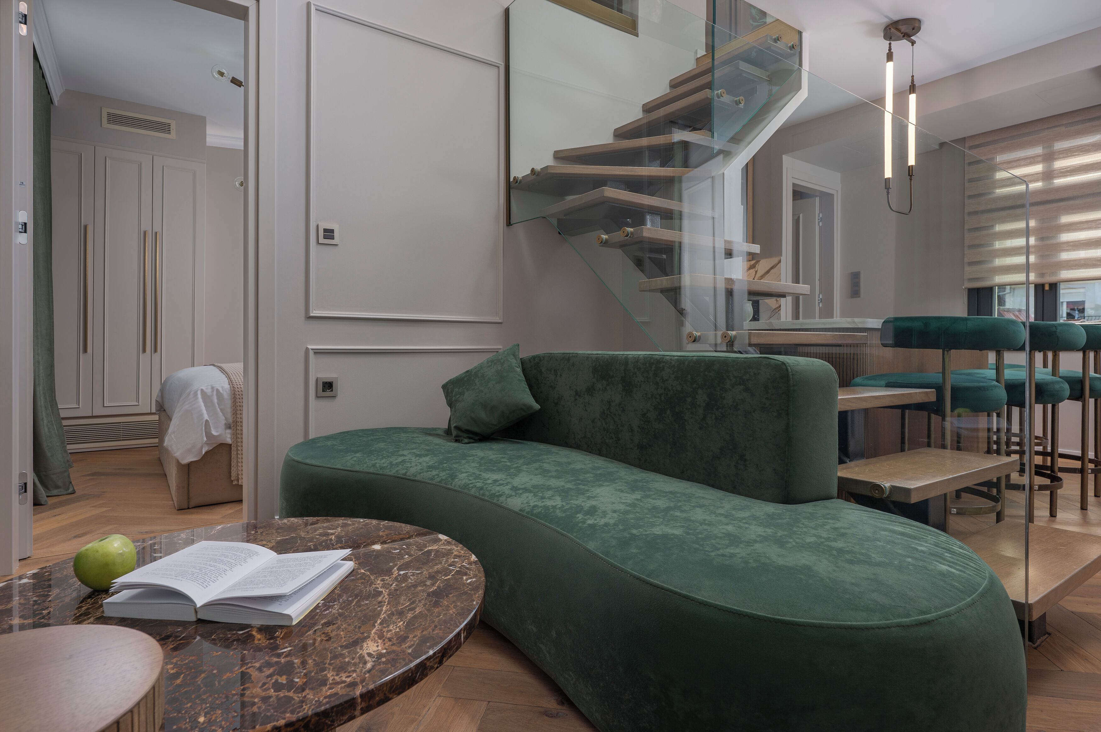Un espacio de estar moderno con un sofá verde curvo, una mesa de centro de mármol y una escalera con barandillas de cristal.
