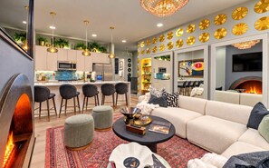 Ein modernes Wohnzimmer mit einer Couch, Hocker und einem Couchtisch. Es gibt eine Küchenzeile mit Barhocker und Pendelleuchten.