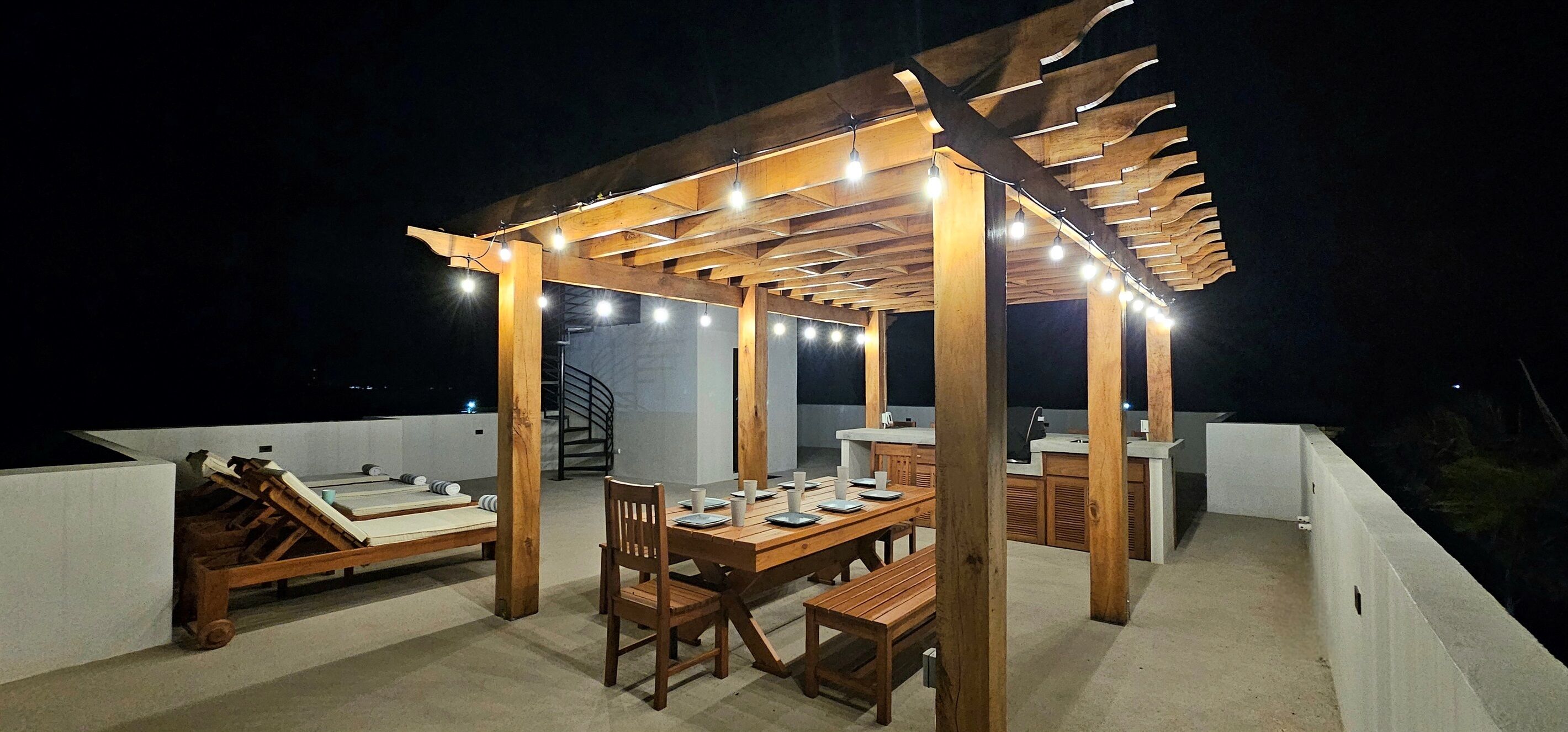 An outdoor patio with a wooden pergola, a dining table with chairs, and a kitchen area.