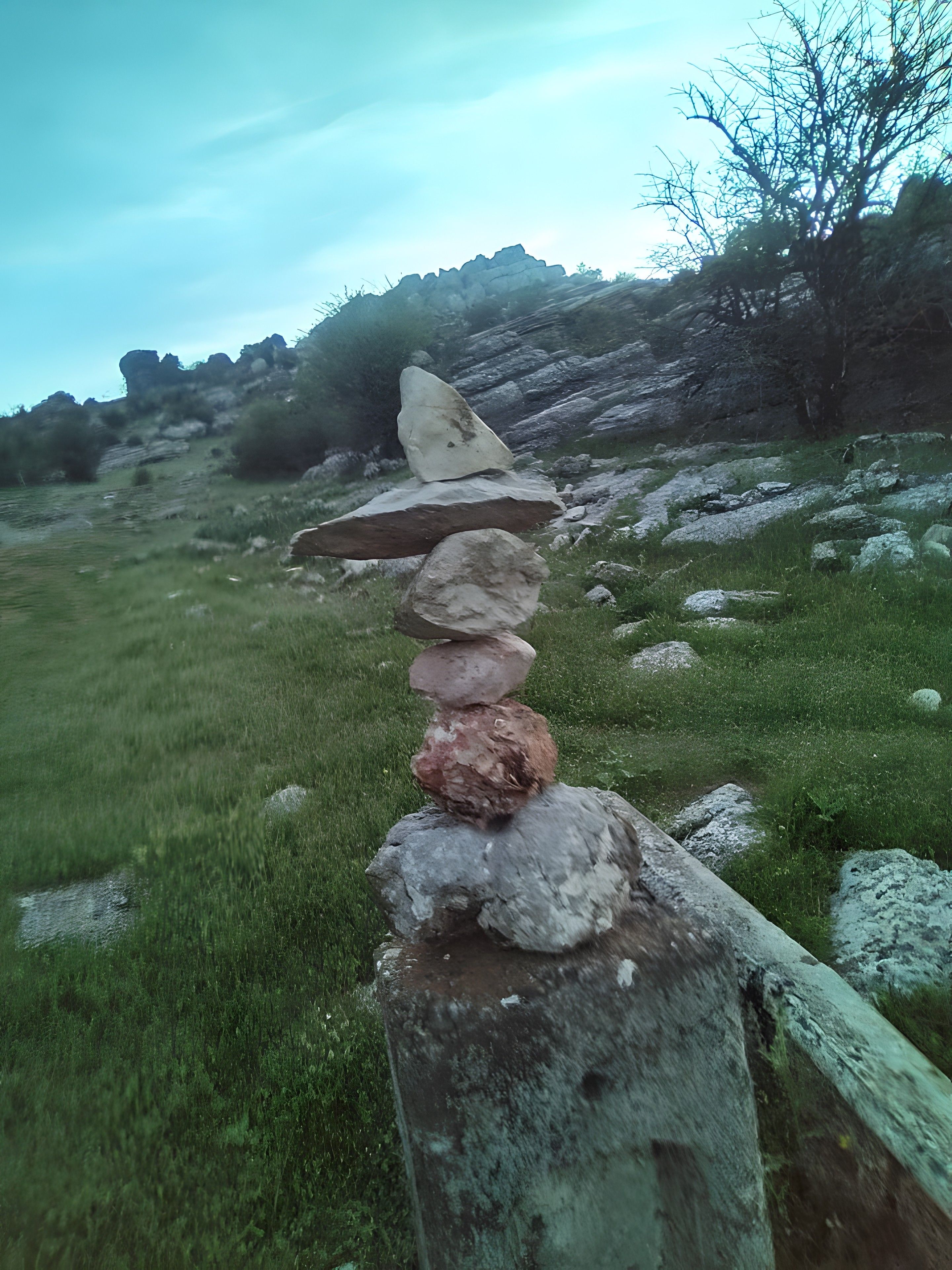Una pila de piedras sobre un poste de madera en un campo cubierto de hierba, con una colina rocosa al fondo.