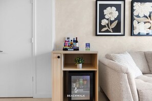 A living room with a wooden shelf holding snacks, a small potted plant, and a refrigerator with beverages. - Elevated Living - 2BR - Intracoastal Views (Hallandale Beach)