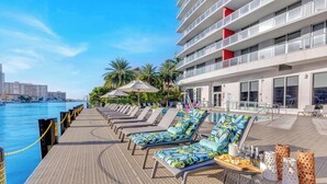 A poolside area with sun loungers and umbrellas, a dining table with snacks, and a view of the water and city skyline. - Elevated Living - 2BR - Intracoastal Views (Hallandale Beach)