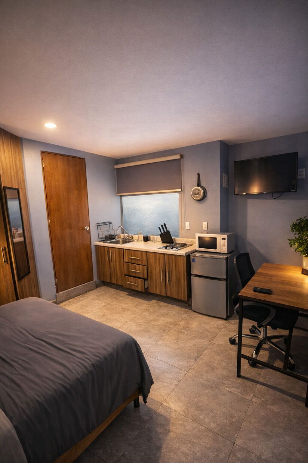 A compact room with a bed, kitchenette, and a desk with a chair.