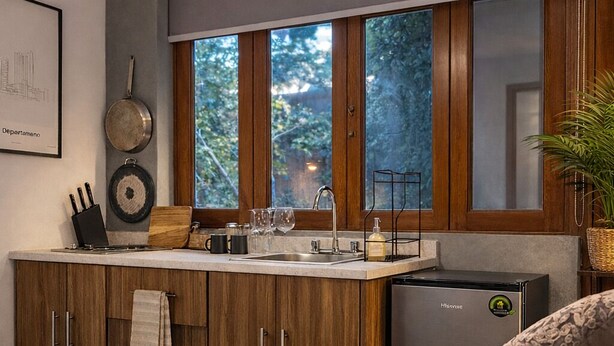 A modern bedroom with a large bed, a small kitchenette with a refrigerator, and a window with a view of trees. - Bowra Orquideas (Zapopan)