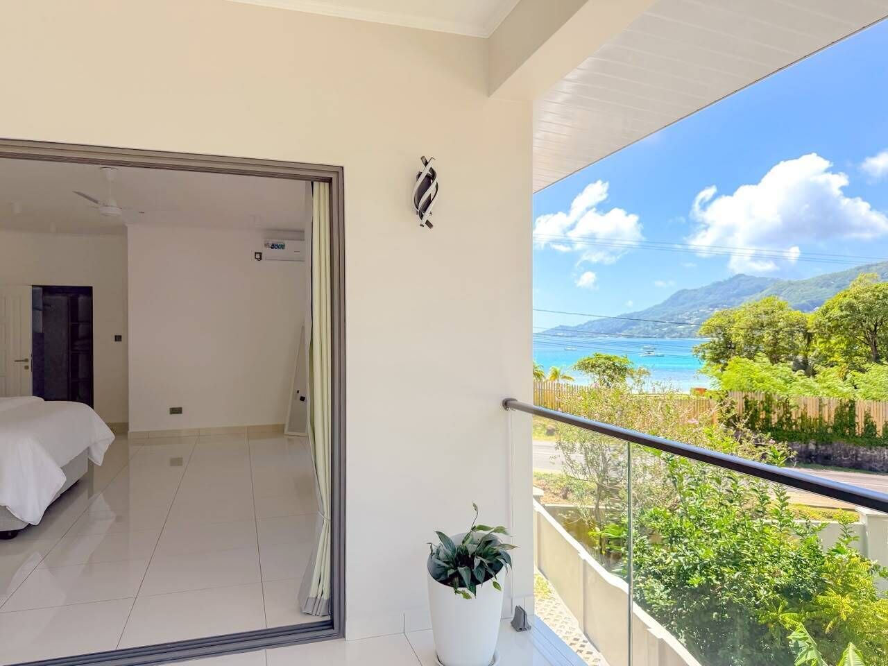 Un balcone con vista sul paesaggio costiero, un letto nella stanza adiacente e una pianta in vaso sul balcone.