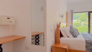 A bedroom with a bed, a bedside table, a mirror, and a window with a view of greenery.