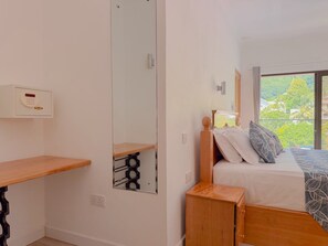 A bedroom with a bed, a bedside table, a mirror, and a window with a view of greenery. - Cove Holiday Apartments (Mahe Island)