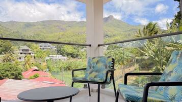 Un balcón con dos sillas y una mesita, con vista a un paisaje montañoso con vegetación y edificios.