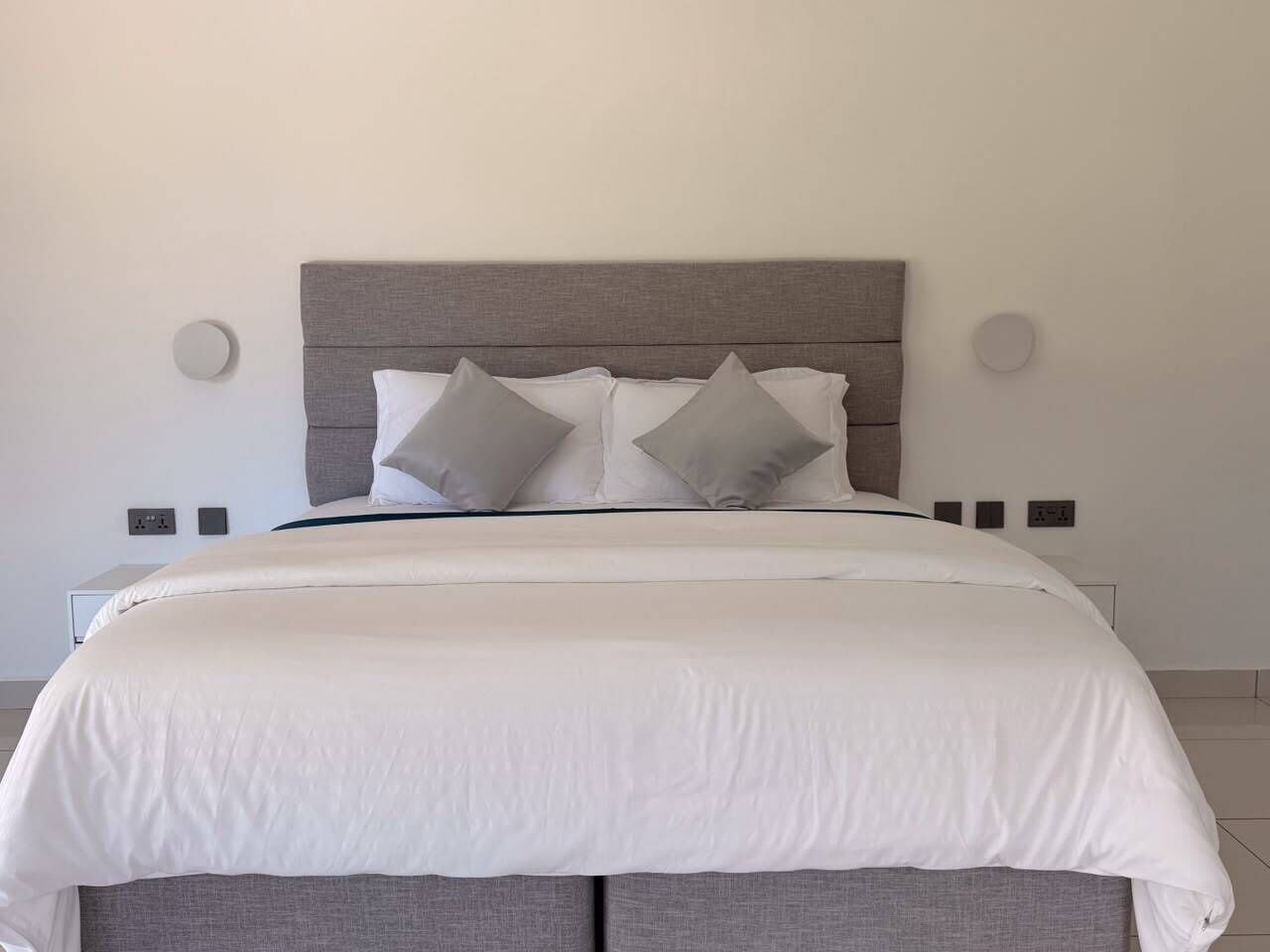 Uma cama com cabeceira cinza, roupa de cama branca e duas mesas de cabeceira com abajures.