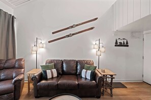 Ein Wohnzimmer mit braunem Ledersofa, zwei Stehlampen, einem kleinen Holztisch und einer Wanddekoration zum Thema Skifahren.