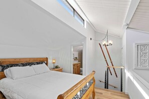 Ein Schlafzimmer mit einem hölzernen Bett, einem Nachttisch, einer Lampe, einer Chandelieber und Blick auf eine Küche durch eine offene Tür.