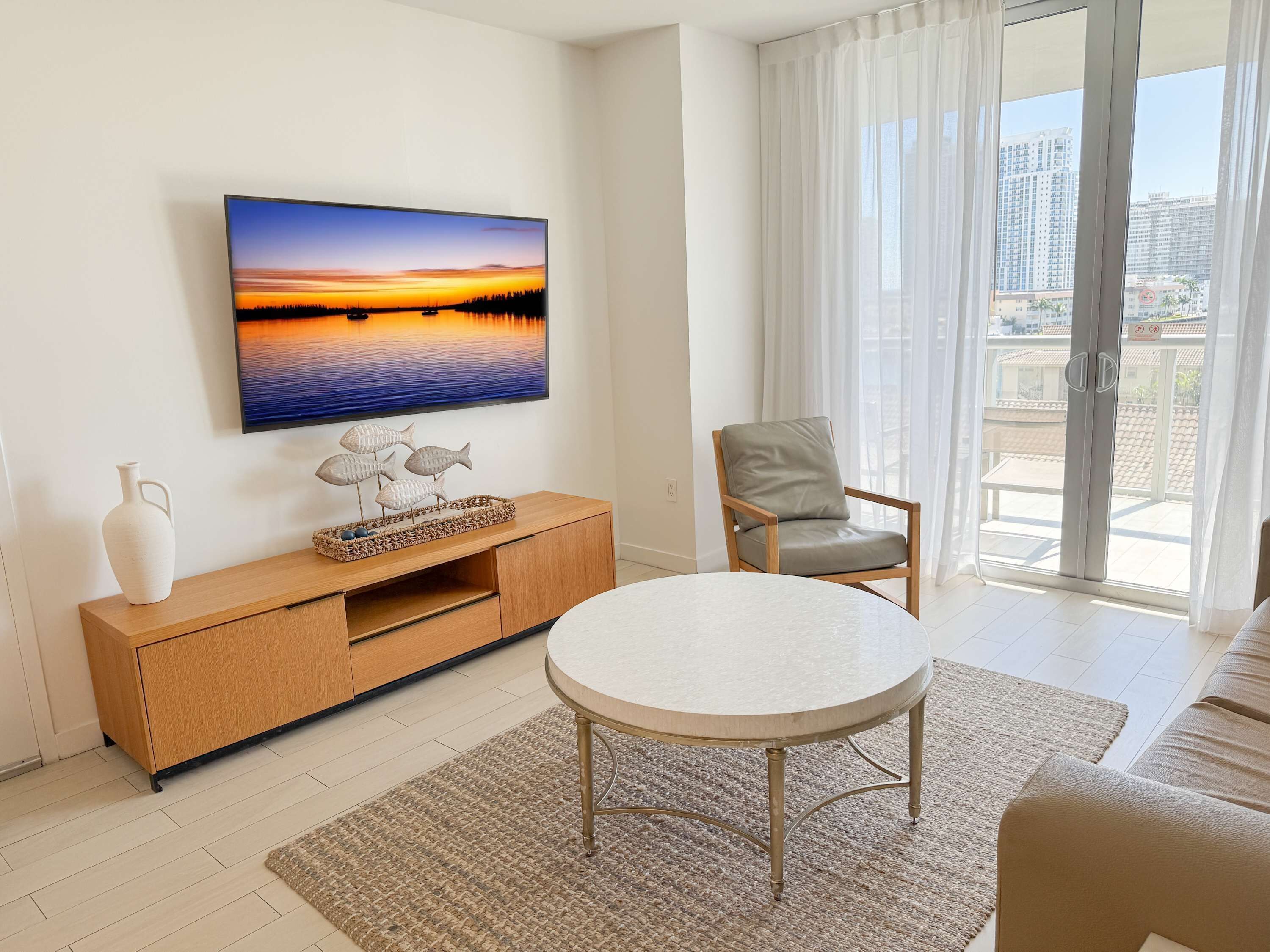 A modern living room with a flat-screen TV, a wooden cabinet, a round table, a grey chair, and a view of a cityscape through a sliding glass door.