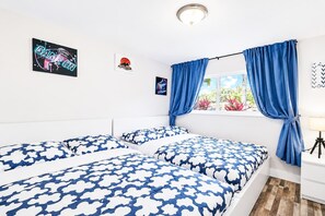 Une chambre avec deux lits, une fenêtre avec des rideaux, un mur décoré d’œuvres d’art et une table de chevet avec une lampe.