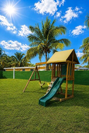 Un terrain de jeux avec des balançoires et un toboggan, entouré de pelouse et de palmiers.