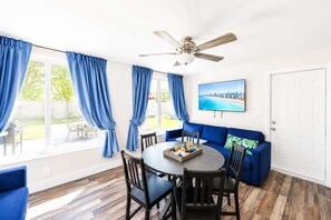 Ein Zimmer mit Esstisch, Stühlen, einem blauen Sofa, einem Deckenventilator und einem an der Wand befestigten Fernseher.
