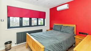 Un dormitorio con cama, ventana con persiana roja, radiador, mesita de noche de madera y unidad de aire acondicionado.