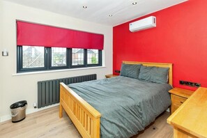 A bedroom with a bed, a window with a red blind, a radiator, a wooden nightstand, and an air conditioning unit.