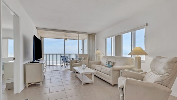 A spacious living room with a balcony, a flat-screen TV, a sofa, a coffee table, and a lamp. - Gulf-Front Bonita Beach, FL Condo -  Private Beach Access + Pool / Gulf views (Bonita Springs)