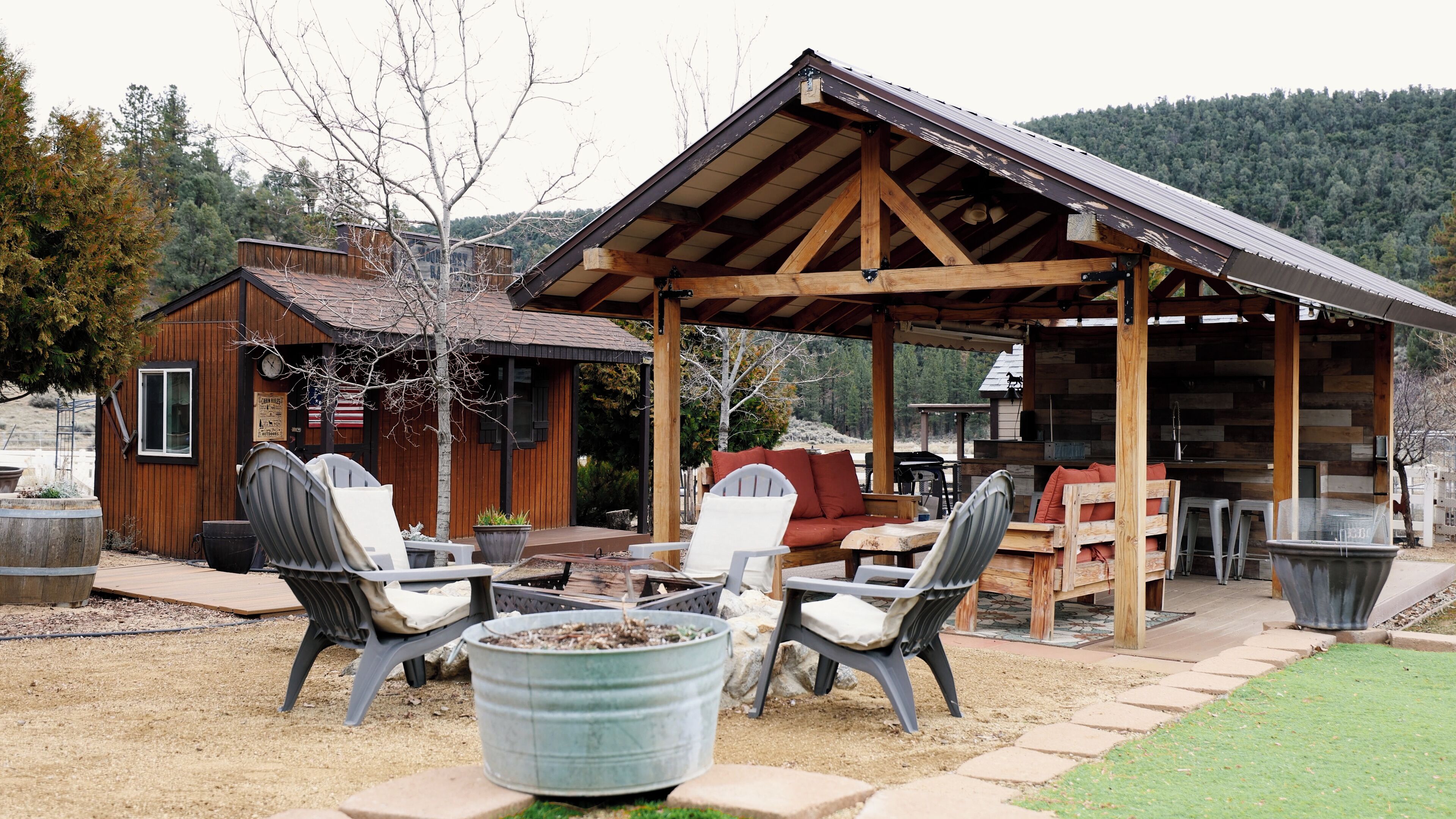 A rustic outdoor seating area with Adirondack chairs, a fire pit, and a wooden structure with a metal roof.