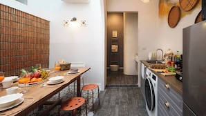 A compact kitchen with a washing machine, a dining area with a table set for two, and a small bathroom visible in the background. - The Post and Barber · Cook In, Hang Out, Sleep Eight (Bang Rak)