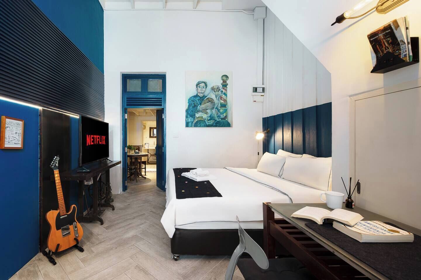 A modern hotel room with a bed, a desk, a guitar, a TV, and a painting on the wall.