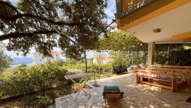 A patio with a table and bench, surrounded by greenery and a view of a building. - One bedroom apartment with sea view (1) (Loviste)