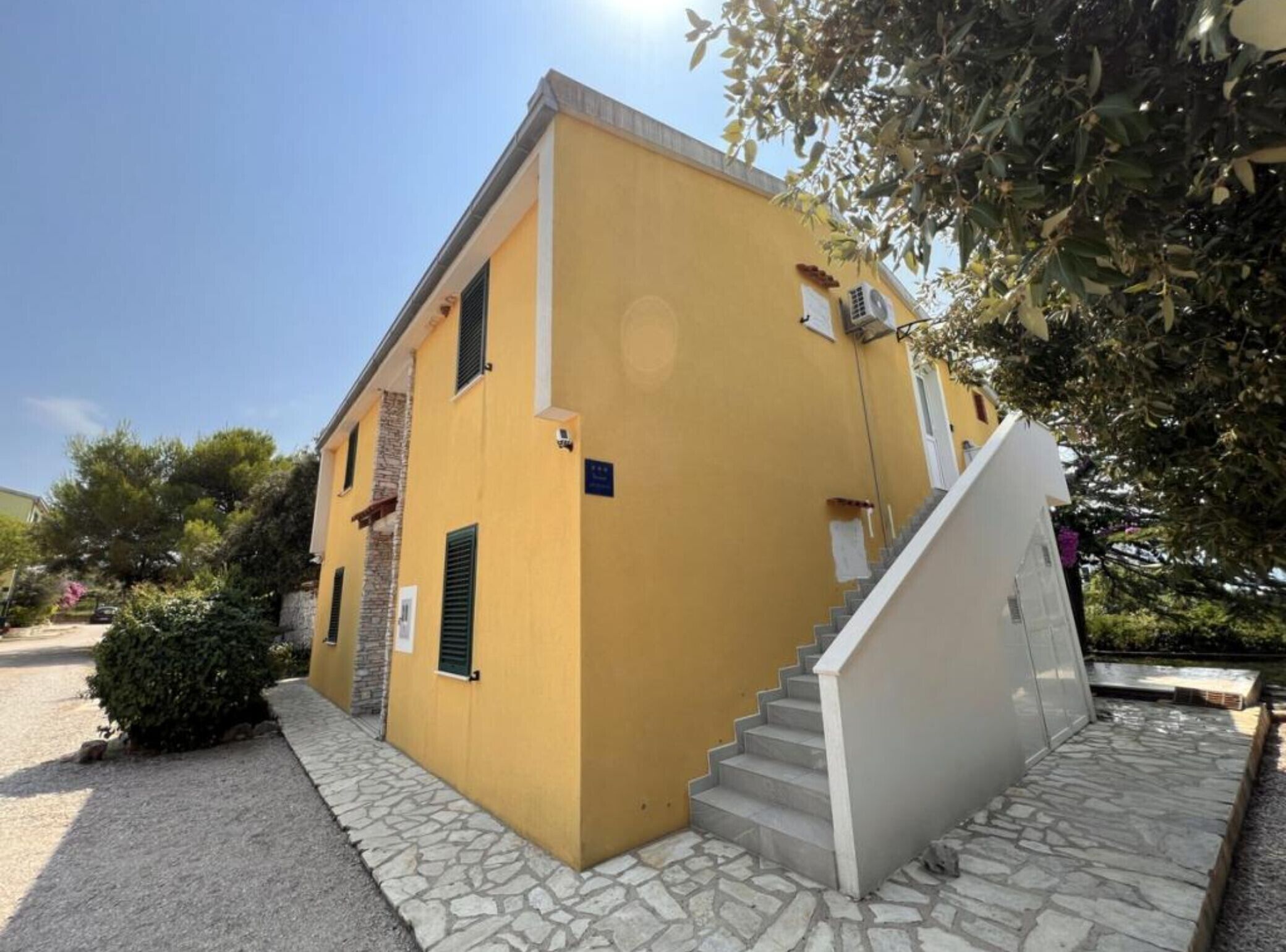 A yellow building with a staircase leading to the entrance, surrounded by greenery.