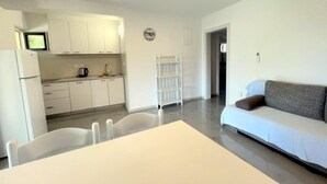 A modern kitchen with white cabinets, a dining table with chairs, a sofa, and a white shelf unit. - One bedroom apartment with sea view (1) (Loviste)
