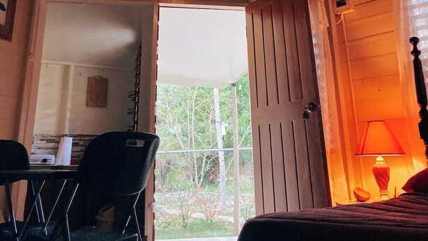 A room with a thatched roof, a bed, a desk with a chair, and a view of the outdoors.