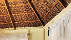 A room with a thatched ceiling, a black leather sofa, and white curtains.