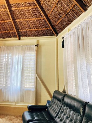 A room with a thatched ceiling, a black leather sofa, and white curtains.