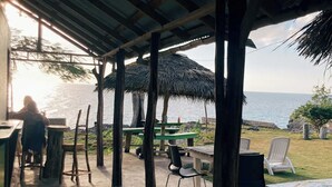 An outdoor seating area with tables and chairs, a thatched roof, and a view of the sea.
