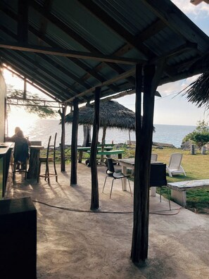 An outdoor seating area with tables and chairs, a thatched roof, and a view of the sea.