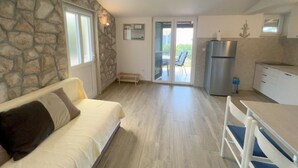 Une pièce avec un mur en pierre, un canapé blanc, une table à manger avec des chaises, un coin cuisine et une porte-fenêtre coulissante donnant sur un espace extérieur.