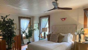 Ein Schlafzimmer mit einem großen Bett, einem Deckenventilator und zwei mit Vorhängen versehenen Fenstern.