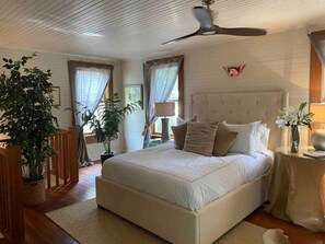 Ein Schlafzimmer mit einem großen Bett, einem Deckenventilator und zwei mit Vorhängen versehenen Fenstern.