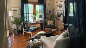 Ein Wohnzimmer mit einer Couch, zwei Sesseln, einem Couchtisch, einem Fernseher und einer Lampe. Es gibt Pflanzen, Vorhänge und Kunstwerke an den Wänden.
