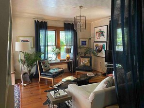Ein Wohnzimmer mit einer Couch, zwei Sesseln, einem Couchtisch, einem Fernseher und einer Lampe. Es gibt Pflanzen, Vorhänge und Kunstwerke an den Wänden.