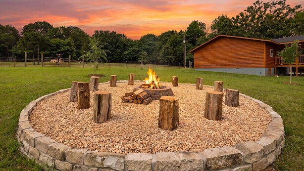 A circular fire pit with logs, surrounded by a stone border, set in a grassy area with a wooden building in the background. - Cloud Heaven Lodge. Close to Eureka Springs, 5 minutes to Eagle Rock Marina/TRL. (Holiday Island)