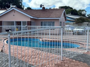 Una piscina con valla blanca, patio de ladrillo y una casa con techo de tejas al fondo.